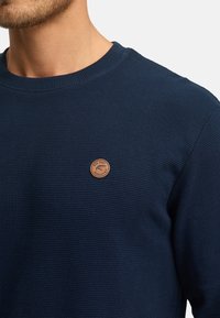 Marineblå ribbet sweater med rund halsudskæring og en lille cirkulær læderlogo-patch på brystet. Blød tekstur og tætsiddende design.