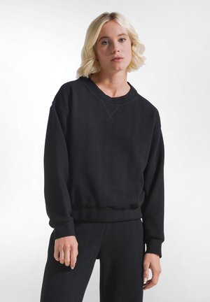 Lyshåret kvinde iført en sort struktureret sweatshirt med lange ærmer og matchende bukser, stående mod en ensfarvet hvid baggrund.