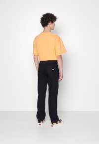 Vaqueros negros de corte recto, camiseta naranja clara y zapatillas coloridas con estampado, vistas desde la parte de atrás contra un fondo liso.