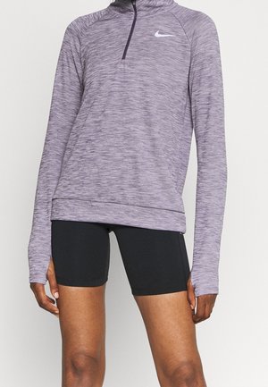 Personne portant un haut de sport violet à manches longues avec des trous pour les pouces et un short noir ajusté, debout devant un fond clair uni.