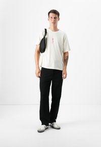 Young man stands confidently, wearing a light shirt with a rose design, black pants, and sneakers, with a black bag slung over his shoulder.