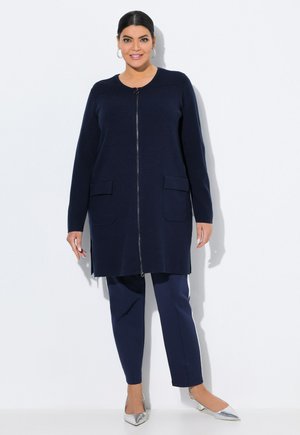 Cardigan long marine avec fermeture éclair à l'avant, deux poches avant et texture côtelée. Assorti à un pantalon marine et des chaussures argentées à bout pointu.