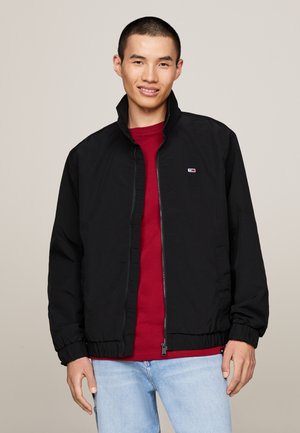 Chaqueta negra con cremallera, cuello alto, puños elásticos y dobladillo. Llevada sobre una camisa roja. Presenta un pequeño logo en el lado izquierdo del pecho.