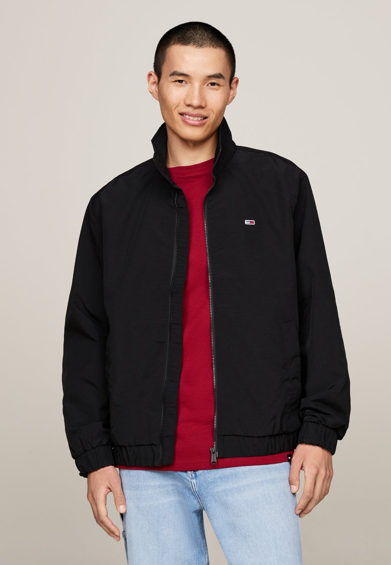 Zwarte ritsjack met staande kraag, elastische manchetten en onderrand. Gedragen over een rood shirt. Bevat een klein logo op de linkerborst.