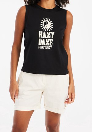 Kobieta nosząca czarny bezrękawnik z białym wzorem słońca i yin-yang oraz napisem „HAZY DAZE PROTEST”, w połączeniu z jasnymi szortami.
