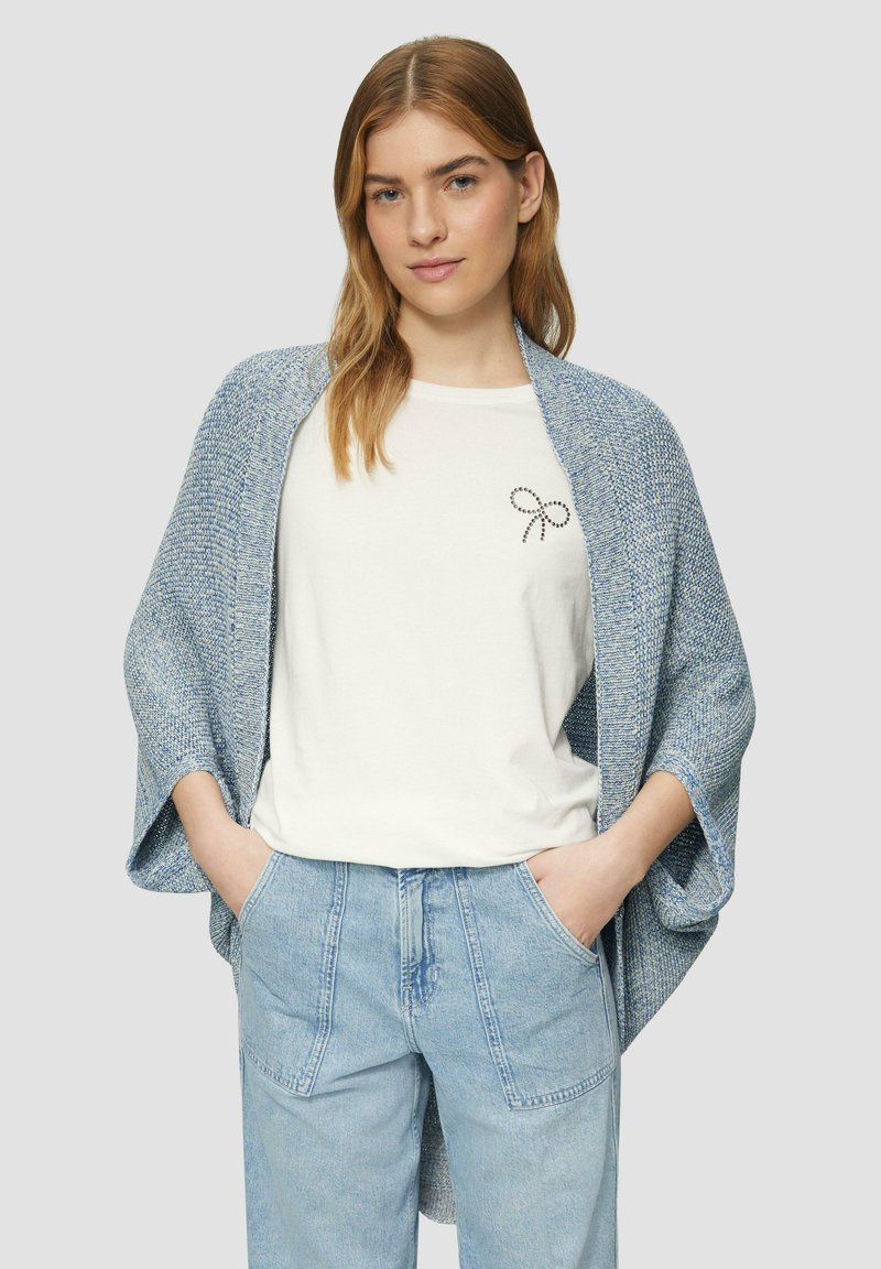 Gros cardigan en tricot bleu clair à devant ouvert, porté sur une chemise pâle avec un motif de nœud à pois, associé à un pantalon en denim bleu clair.