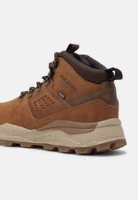 Brauner Wanderschuh aus Leder mit gepolstertem Kragen, strukturiertem beigem подошва und Metallösen. Mit Logo und perforierter Detailierung an der Seite.