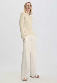 Beige gerippter Pullover mit langen Ärmeln, kombiniert mit lockeren weißen Hosen und beigen Sandalen. Weicher Stoff und entspannter Schnitt.