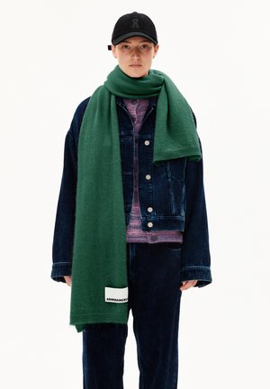 Persoon in een zwarte pet, een donkerblauwe corduroy jas en broek, een paars shirt en een lange groene sjaal met merklabel, staand tegen een witte achtergrond.