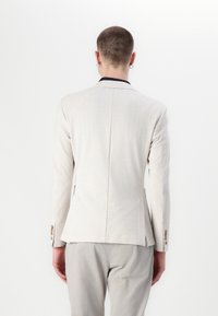Lys grå blazer med en tettsittende fasong, strukturerte skuldre og to lommer. Har en marineblå krage og knappeakenter på ermene.