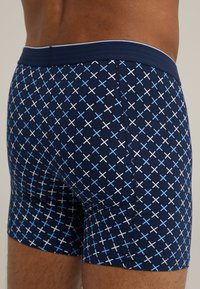 Navy kék boxer alsónadrág geometrikus mintázattal, amely keresztbe fekvő fehér és világoskék X formákat tartalmaz. Rugalmas derékpánt és sima szövettextúra.