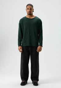 Groen shirt met lange mouwen met versleten details, gecombineerd met zwarte broek met wijde pijpen en zwarte schoenen met roze veters. Effen achtergrond.