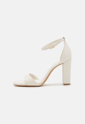 Sandale blanche à talons hauts avec une bride de cheville, dotée de deux brides avant, d'une finition en cuir lisse et d'un talon bloc avec une semelle marron clair.