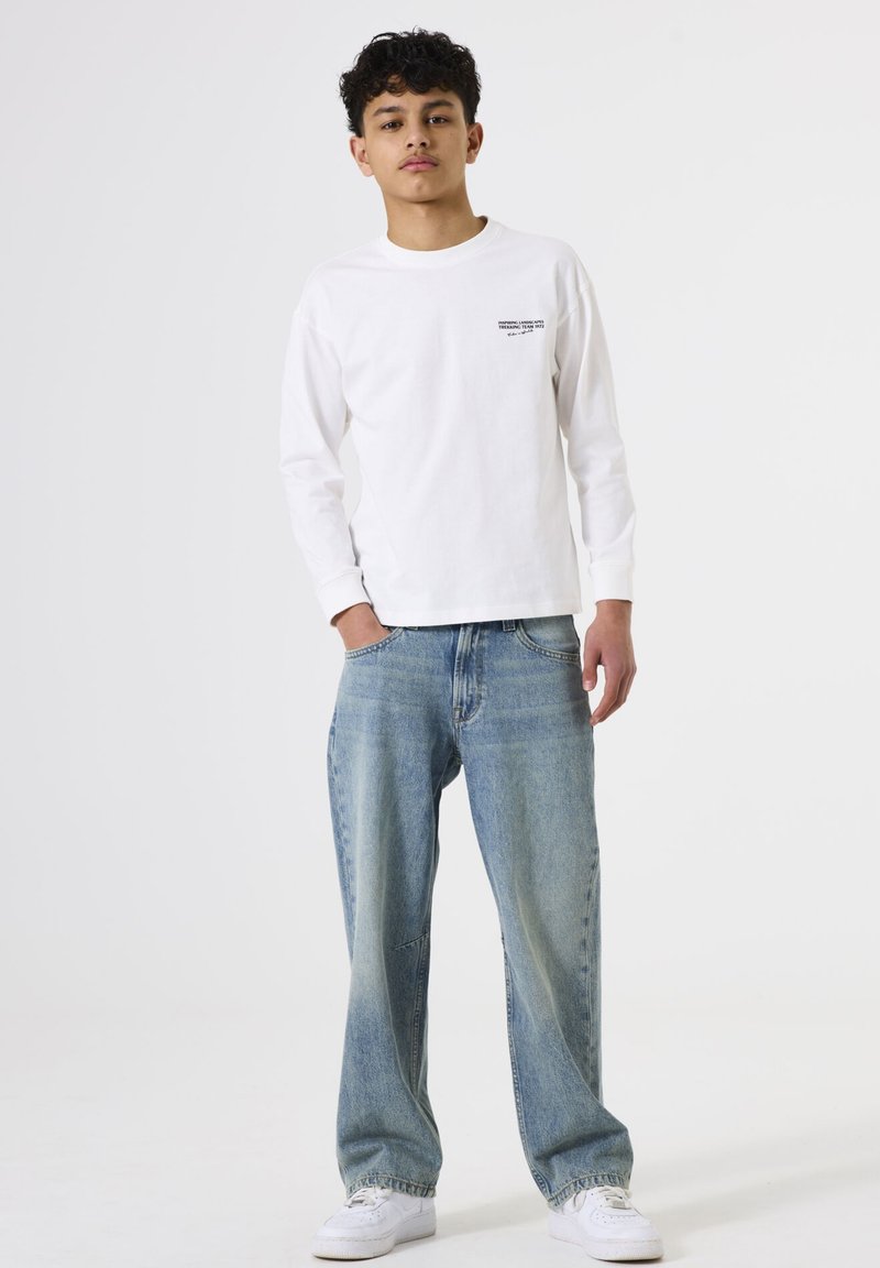 Wit katoenen T-shirt met lange mouwen en bedrukte tekst op de linkerborst, gecombineerd met losse lichtblauwe spijkerbroek en witte sneakers.