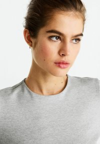 Sivá tričko s krátkym rukávom a okrúhlym výstrihom, hladká textúra a minimalistický dizajn. Modelka má svetlú pleť a prírodný makeup.