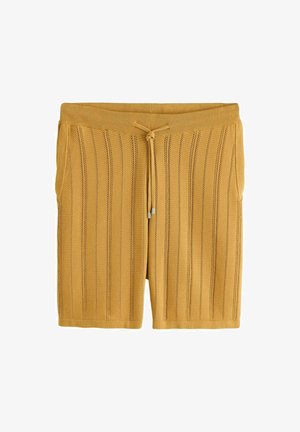 Tânăr îmbrăcat în pantaloni scurți tricotati de culoare muștar și tricou polo, purtând sandale negre și ochelari de soare, sprijinit de un zid de piatră lângă plante verzi.