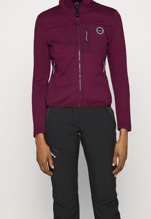 Mujer con chaqueta ajustada morado oscuro con cremallera, con bolsillos en el pecho y a los lados, combinada con pantalones negros para exteriores.