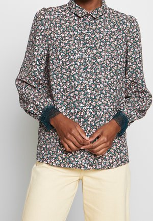 Person iført en langærmet blomstret bluse med pink og hvide blomster, blonderkanter på ærmerne, kombineret med lyse beige bukser.