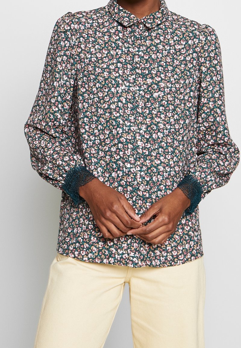 Personne portant un chemisier à manches longues à fleurs roses et blanches, avec des poignets bordés de dentelle, associé à un pantalon beige clair.