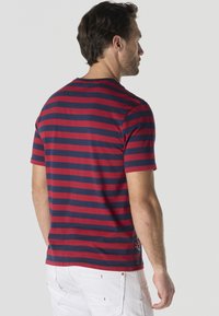 Homme vu de dos portant un t-shirt à manches courtes à rayures horizontales rouges et bleu marine et un pantalon blanc sur un fond uni.