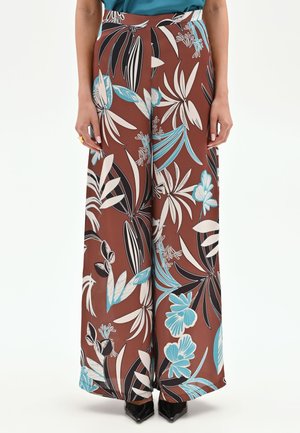 Pantalon marron à jambes larges avec un grand imprimé feuilles tropicales blanches, noires et bleues, porté avec des chaussures noires à bouts pointus et un top bleu sarcelle rentré.