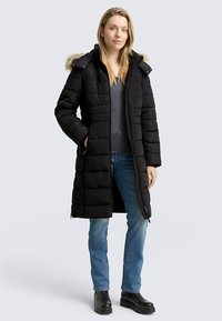 Black padded coat with fur-lined hood, zip closure, and side pockets. Worn over a grey top and blue jeans, displayed on a neutral background.