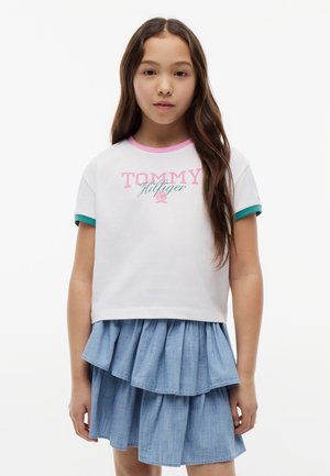 Fille aux longs cheveux bruns portant un t-shirt blanc Tommy Hilfiger avec des bordures roses et vertes et une jupe bleue superposée, debout sur un fond uni.