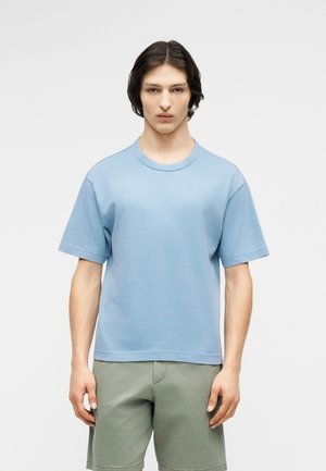 Młoda osoba o średniej długości ciemnych włosach, ubrana w jasno niebieską koszulkę z krótkim rękawem i oliwkowo zielone szorty, stojąca na jednolitym tle.