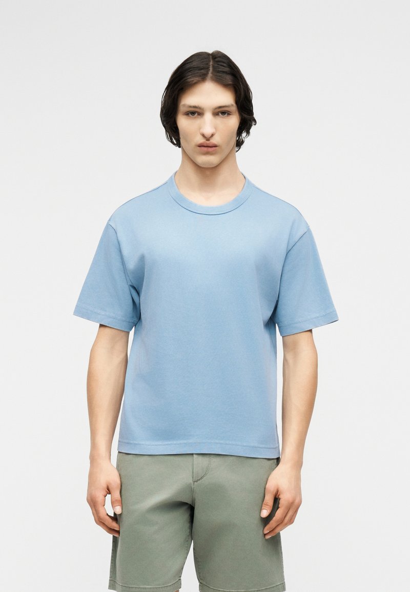 Νεαρό άτομο με μεσαίου μήκους σκούρα μαλλιά, φορεμένη ανοιχτό μπλε κοντομάνικη μπλούζα και χακί σορτς, που στέκεται μπροστά σε απλό φόντο.