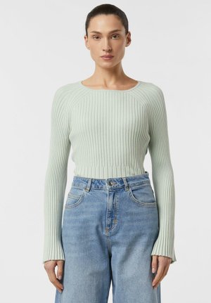 Kobieta z ciemnymi włosami związanymi do tyłu, ubrana w jasnozielony, prążkowany sweter z długim rękawem i wysokim stanem niebieskich dżinsów na jednolitym tle.