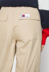 Beige bavlněné kalhoty s hladkým povrchem, které mají dvě zadní kapsy a značkovou etiketu s červenými, bílými a modrými akcenty.