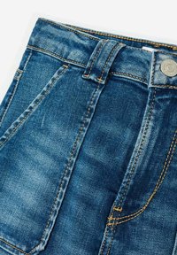 Jean en denim avec un lavage bleu foncé, présentant des surpiqures contrastantes, une fermeture éclair et un bouton en métal. Poches avant et texture légèrement délavée.