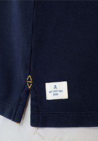 Polo en color azul marino con tejido texturizado, que presenta una etiqueta de marca blanca con un ícono de calavera y detalle de costura amarilla en el dobladillo.