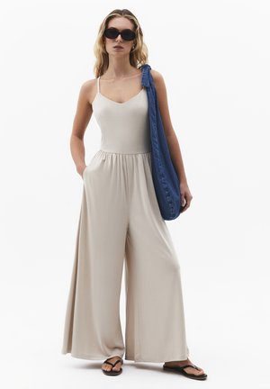 Kvinde iført beige jumpsuit med brede ben, sorte solbriller, sorte flip-flops og bærende en blå skuldertaske på sin højre skulder mod en hvid baggrund.