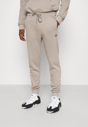 Beige cotton jogger pants with an elastic waistband, drawstring, and side pockets. Black logo detail on the left thigh. Worn with black and white sneakers.