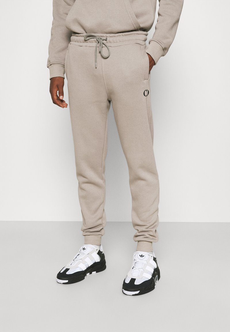 Beige cotton jogger pants with an elastic waistband, drawstring, and side pockets. Black logo detail on the left thigh. Worn with black and white sneakers.