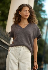 Gray short-sleeve sweater with a v-neck design, paired with high-waisted off-white jeans, held with a hand carrying a patterned bag.