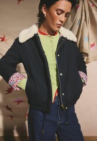 Jachetă bomber bleumarin cu guler din blană artificială crem, cu accente de manșete cu model, purtată peste un top verde cu nasturi și blugi de denim închis la culoare.