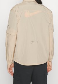 Giacca Nike beige con un tessuto leggero e texturizzato. Presenta un grande logo sul retro, polsini elastici e un leggero motivo stampato.
