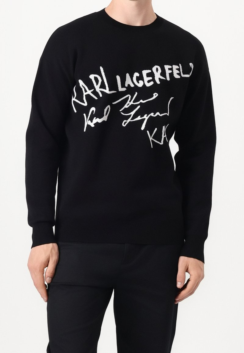 Pull noir à manches longues avec texte et signature blanche en style manuscrit "Karl Lagerfeld" sur la poitrine, porté par une personne debout.