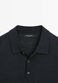 Tmavě námořnická polo košile s ribbovanou strukturou, krátkým límečkem, tříknoflíkovou légou a nenápadnou značkovou štítkou viditelnou blízko výstřihu.