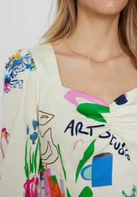 Rochie cu mâneci scurte din țesătură crem, evidențiată de imprimeuri abstracte colorate, cu decolteu pătrat, detalii cu pliuri și accente de colier aurie.