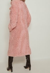 Abrigo rosa esponjoso hecho de tejido texturizado, con un diseño holgado y mangas largas. Combinado con botas negras de tacón alto.