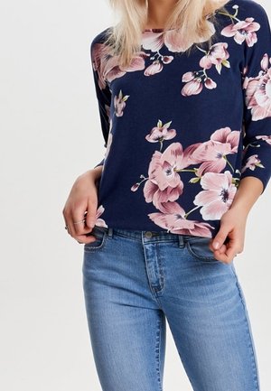 Femme portant une chemise bleu marine à manches longues avec un imprimé floral rose et un jean bleu clair, mains tenant l'ourlet de la chemise.