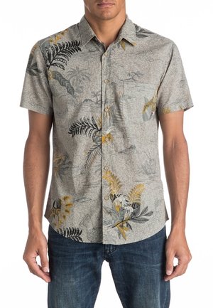 Homme portant une chemise à manches courtes boutonnée avec des motifs de feuilles tropicales et floraux gris, noirs et jaunes, associée à un jean bleu foncé.