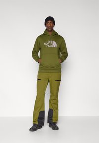 Φούτερ με κουκούλα σε ελαιόχρωμο με λογότυπο, σε συνδυασμό με παντελόνι ελαίου με μαύρες λεπτομέρειες και τσέπες με φερμουάρ. Μαύρα παπούτσια ολοκληρώνουν το ντύσιμο.