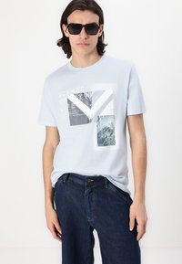 T-shirt à manches courtes bleu clair avec un imprimé graphique comportant des photos superposées et du texte, associé à un jean en denim foncé.