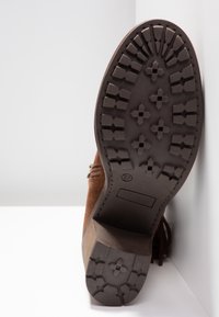 Brązowa zamszowa podeszwa buta z teksturowaną gumową podeszwą z prostokątnymi i krzyżowymi wzorami. Metka pokazuje rozmiar 37.