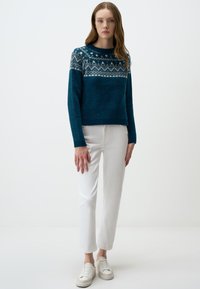 Teal gebreide trui met een patroon in wit en marineblauw, gecombineerd met witte recht gesneden broek en lichte sneakers.
