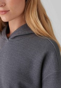 Felpa grigia con cappuccio, in tessuto texturizzato, dalla vestibilità rilassata e scollatura classica. Il tessuto ha una leggera lucentezza, adatta per un look casual.